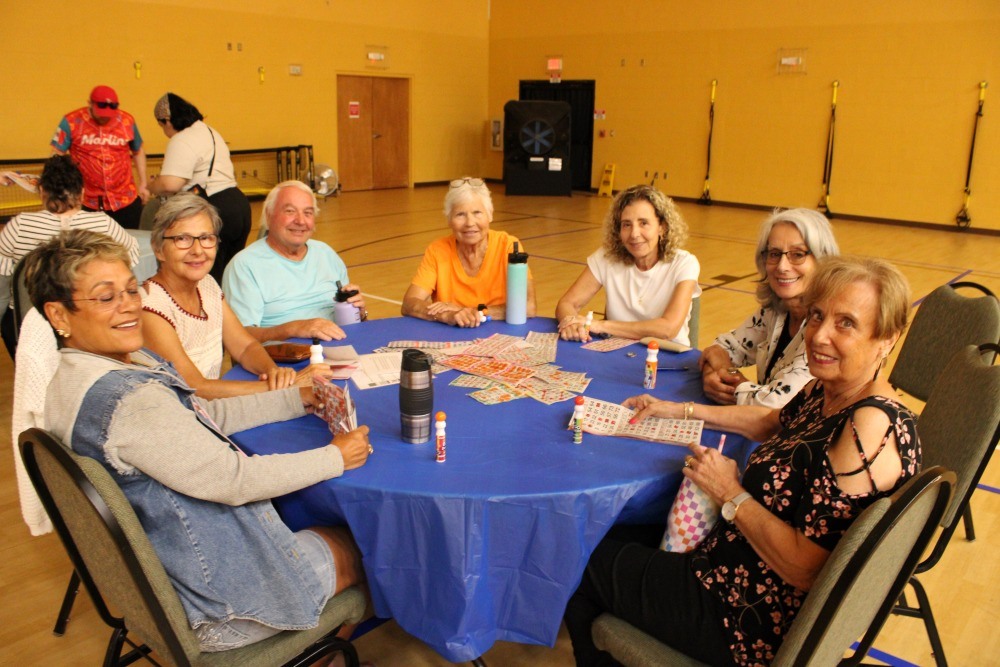 Wide view of the gym showing multiple tables of people playing bingo beneath a large projection screen